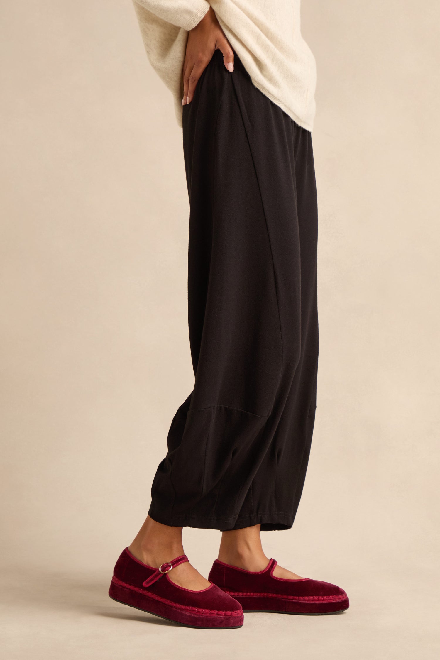 A person wearing the Fluid Crepe Bubble Trouser in loose black with a rounded leg and a cream sweater stands on a beige background, showing burgundy velvet Mary Jane shoes.