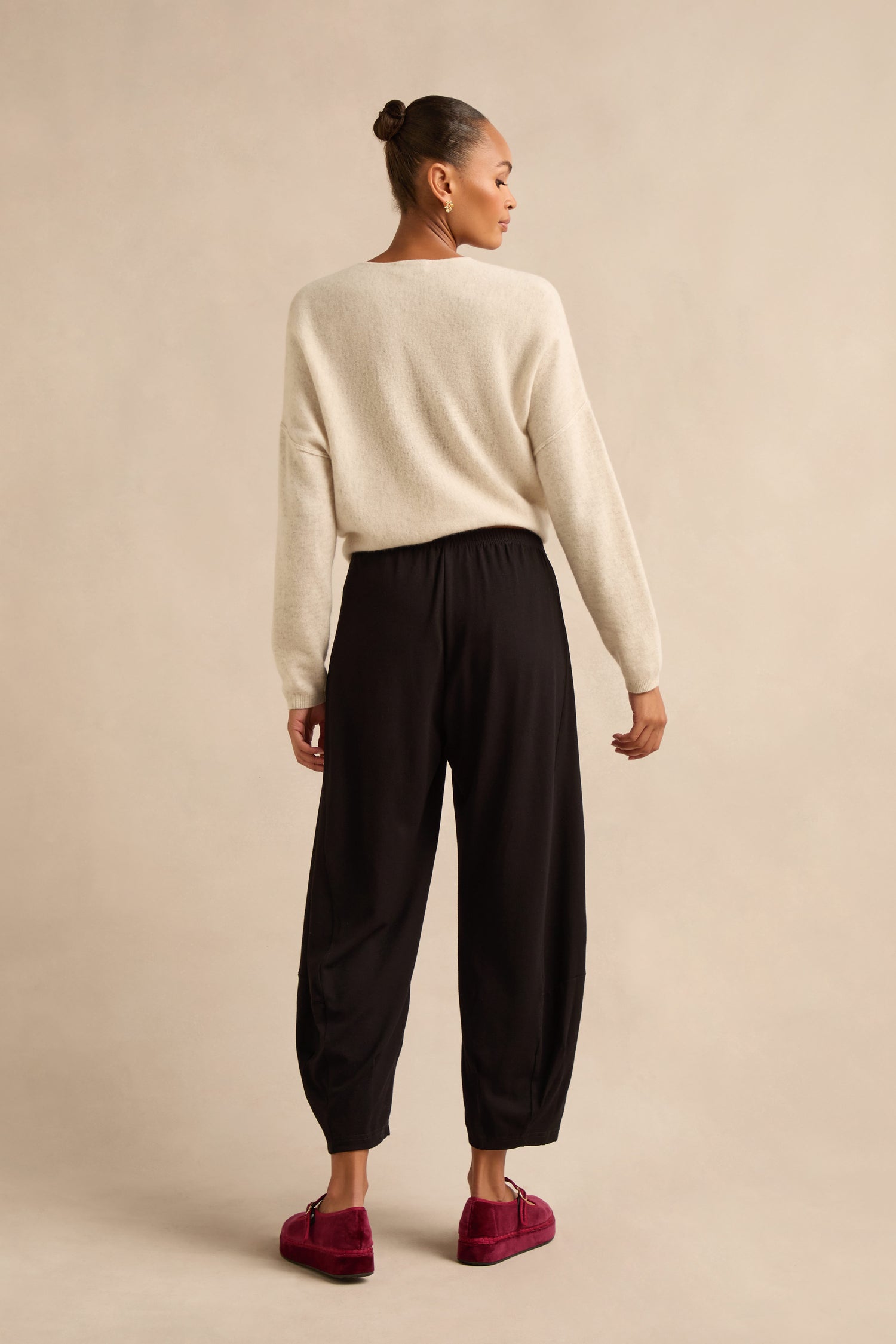 A person stands facing away, wearing a cream sweater, the Fluid Crepe Bubble Trousers in black, and red loafers, against a plain beige background.