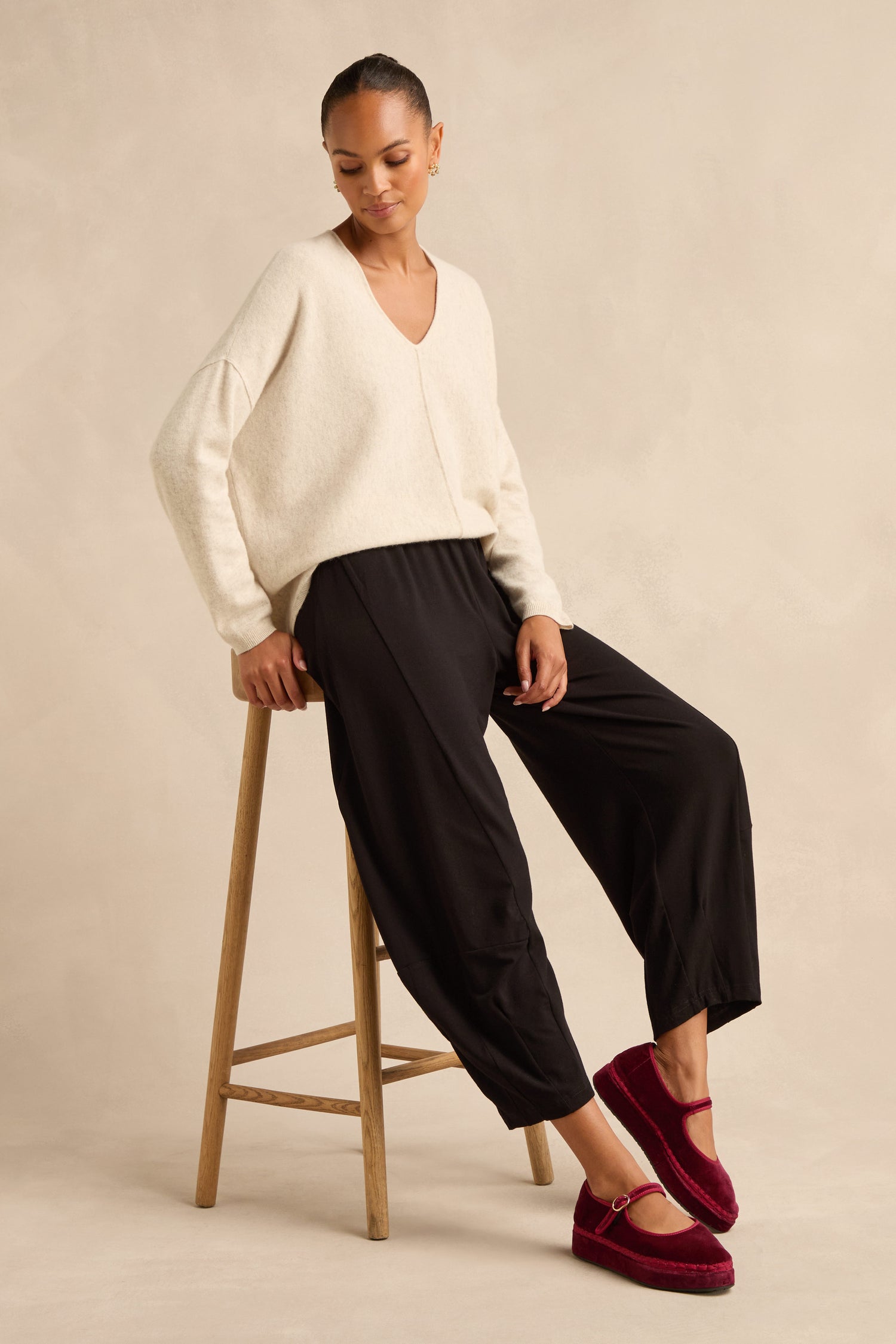 A woman sits on a wooden stool against a neutral background, wearing a cream sweater, burgundy slippers, and the Fluid Crepe Bubble Trouser featuring a slim bubble silhouette and rounded leg.