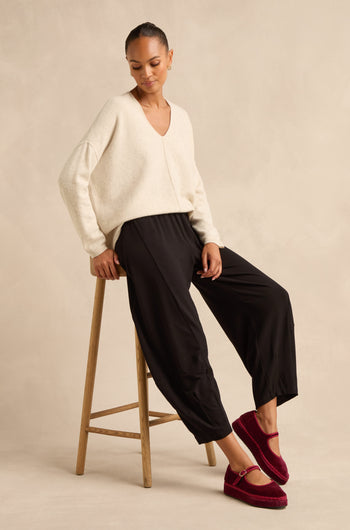 A woman sits on a wooden stool against a neutral background, wearing a cream sweater, burgundy slippers, and the Fluid Crepe Bubble Trouser featuring a slim bubble silhouette and rounded leg.