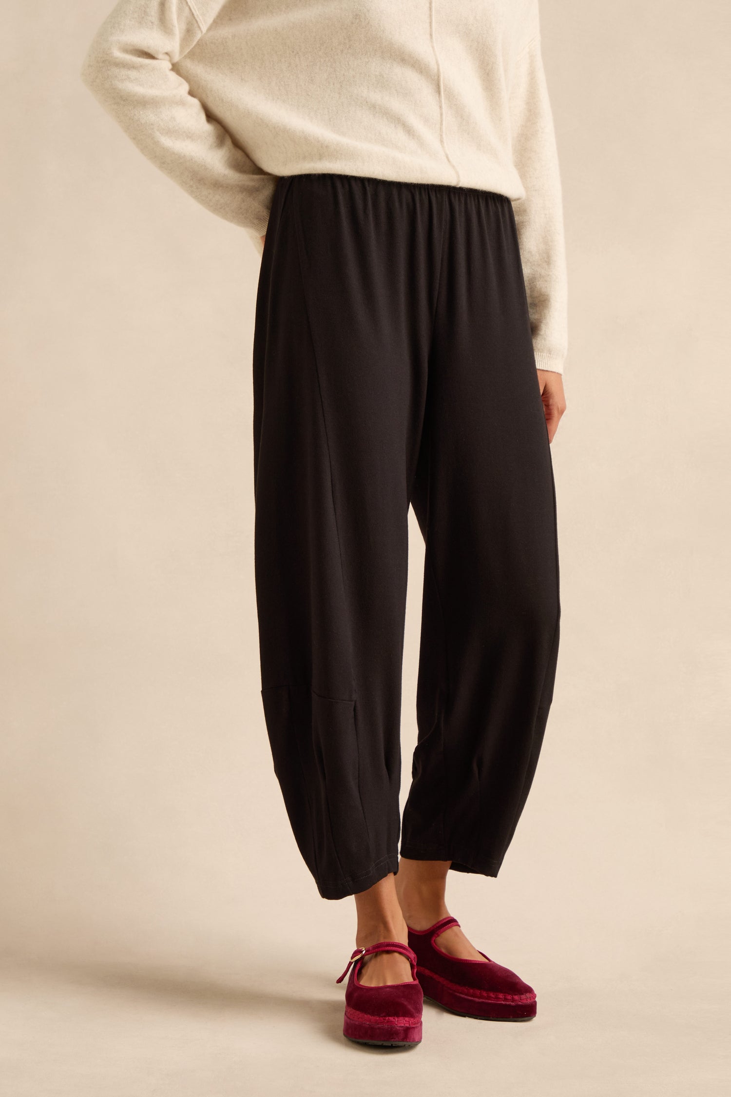 A person wearing a cream sweater, Fluid Crepe Bubble Trouser in black—with a rounded leg and slim bubble silhouette—and red velvet slippers stands against a neutral background.