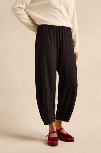 A person wearing a cream sweater, Fluid Crepe Bubble Trouser in black—with a rounded leg and slim bubble silhouette—and red velvet slippers stands against a neutral background.