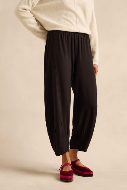 A person wearing a cream sweater, Fluid Crepe Bubble Trouser in black—with a rounded leg and slim bubble silhouette—and red velvet slippers stands against a neutral background.
