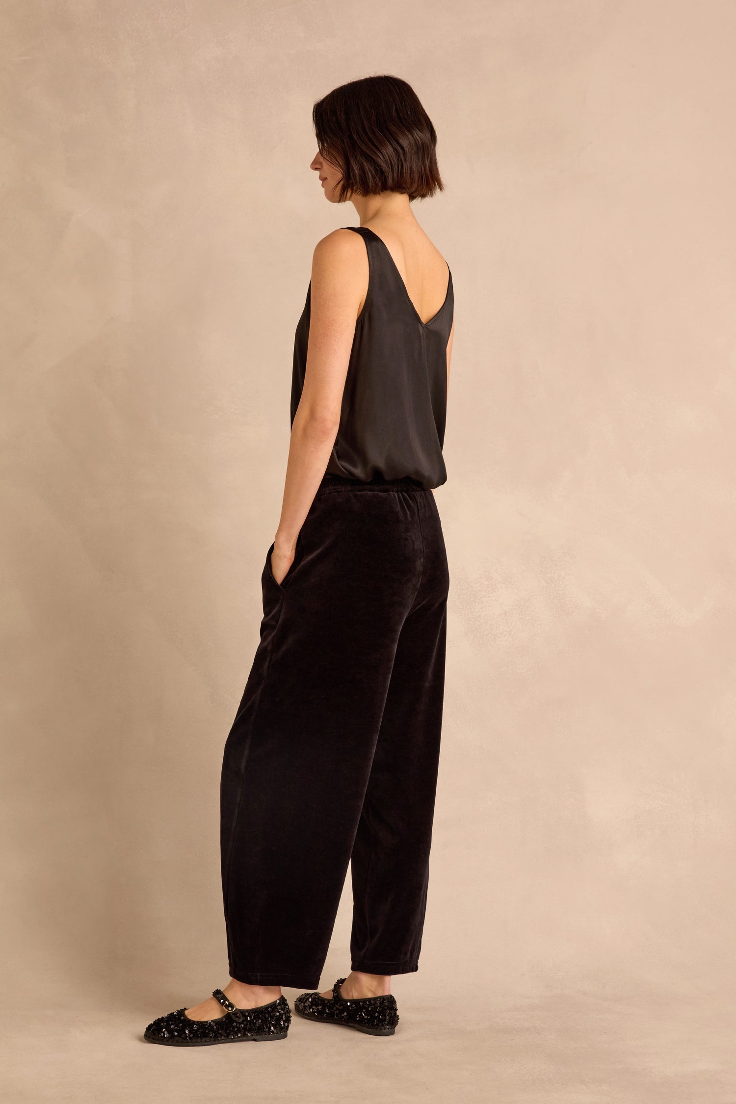 A woman stands sideways against a neutral background, wearing Black Velvet Jersey Bubble Trousers paired with a sleeveless black top and black shoes.