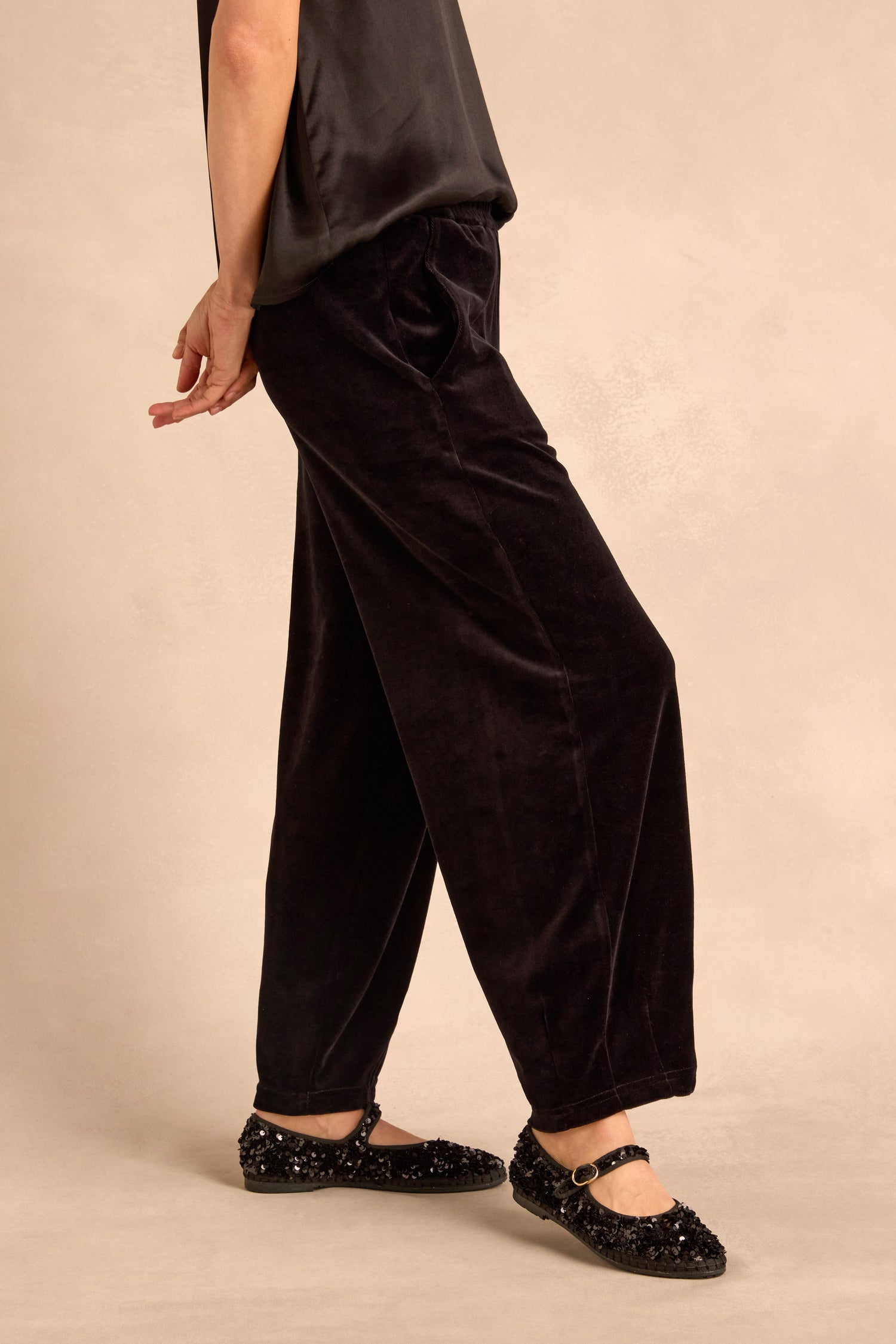 Wearing Black Velvet Jersey Bubble Trousers in a loose bubble silhouette, paired with a dark sleeveless top and black sequined Mary Jane shoes, the person stands against a neutral background.