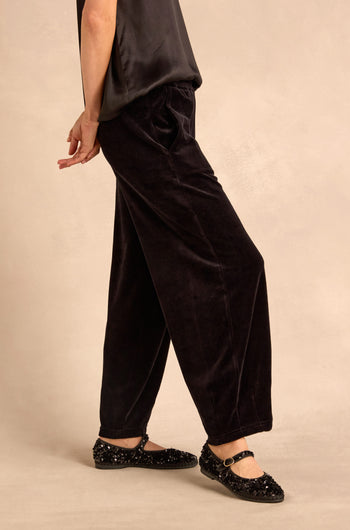 Wearing Black Velvet Jersey Bubble Trousers in a loose bubble silhouette, paired with a dark sleeveless top and black sequined Mary Jane shoes, the person stands against a neutral background.