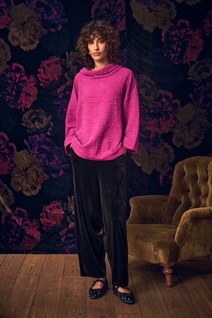 A person wearing a bright pink textured velvet jersey long-sleeved top and Black Velvet Jersey Bubble Trousers stands in front of a floral wallpaper.