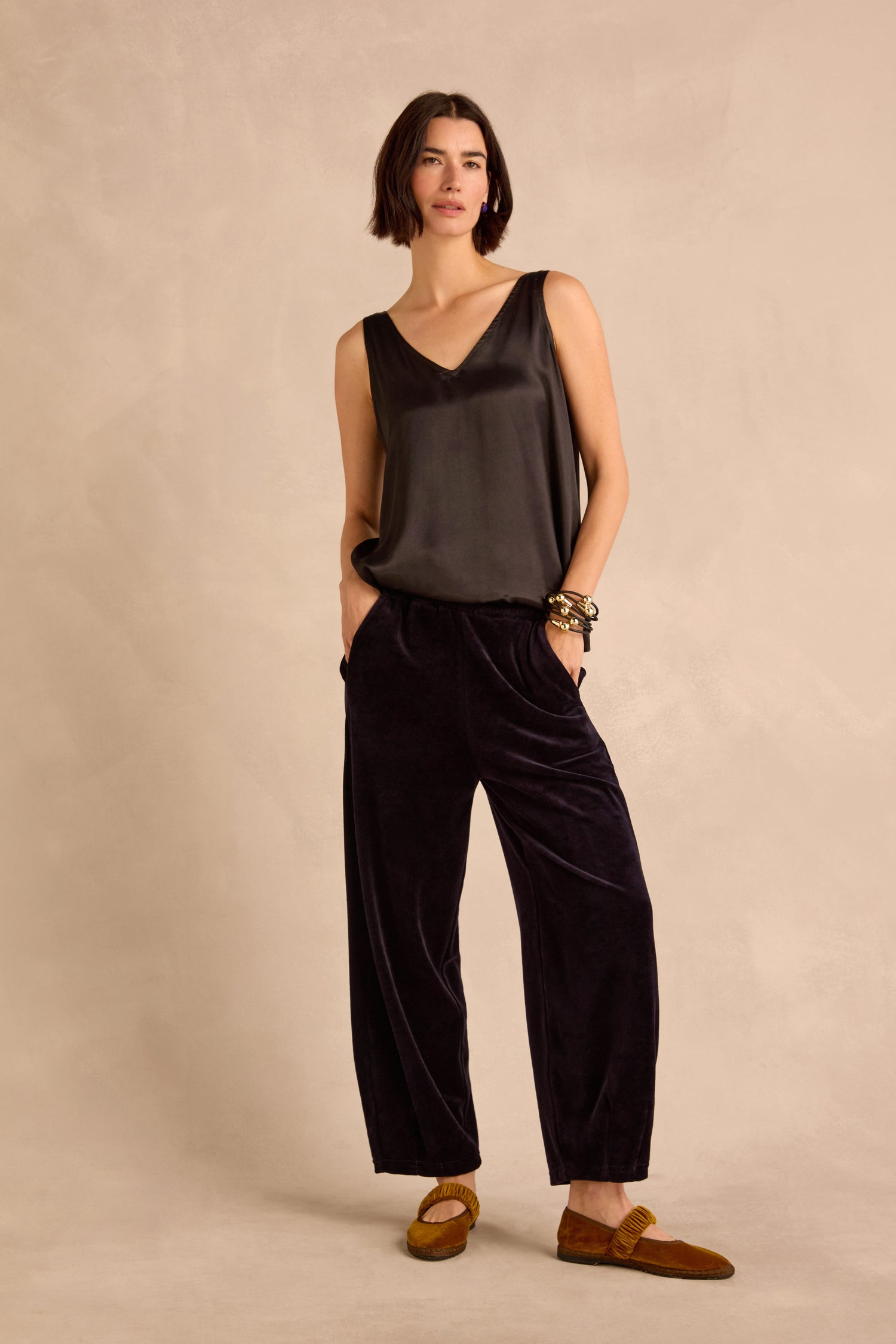 A woman stands against a beige background, hands in her pockets and wearing the effortlessly chic Night Velvet Jersey Bubble Trousers—a loose, bubble-silhouette design crafted in England—paired with a sleeveless black top, tan flats, and bracelets. She maintains a neutral expression.