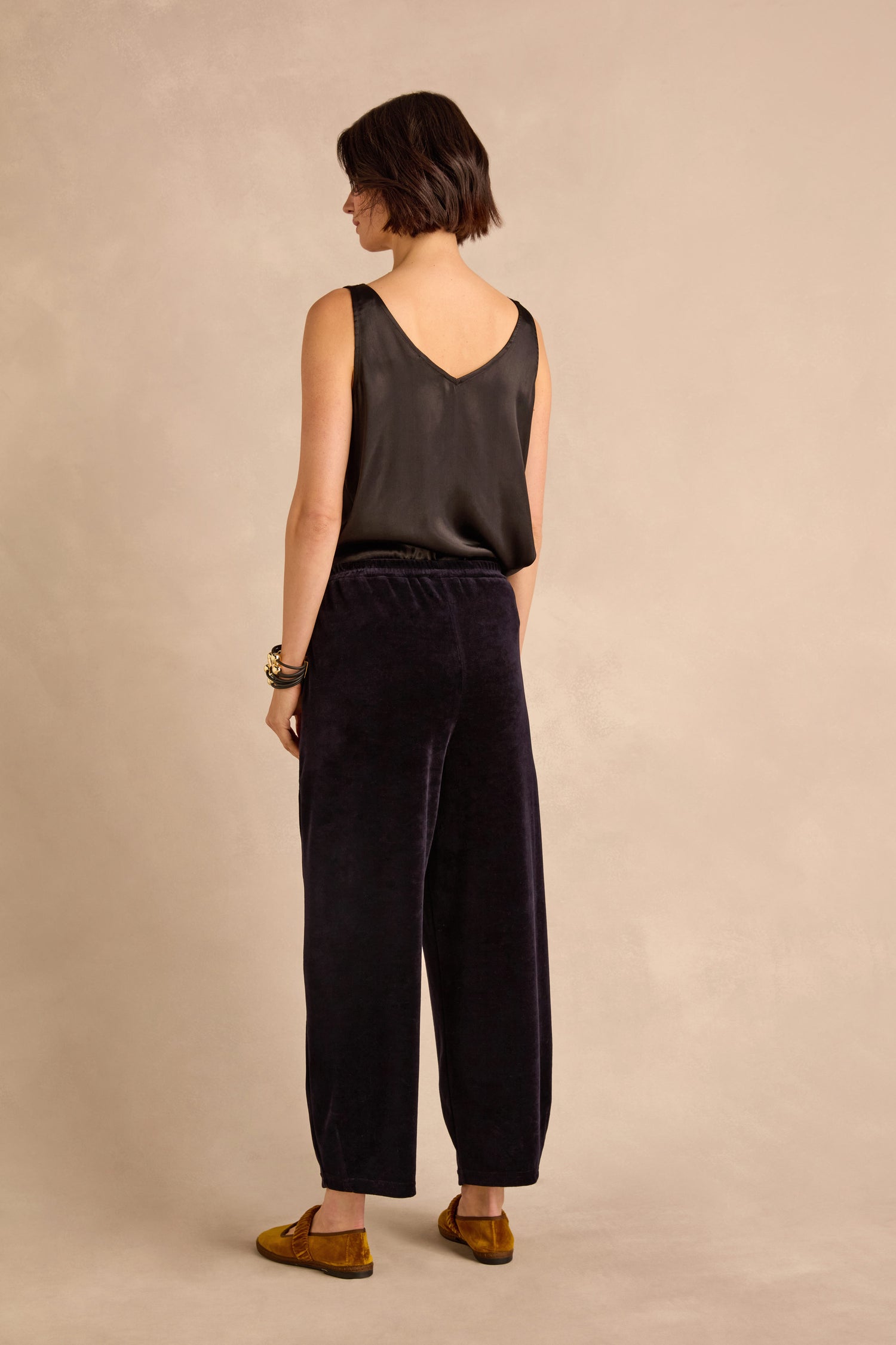 A person stands facing away, wearing a sleeveless black top, the Night Velvet Jersey Bubble Trousers with their subtle bubble silhouette, brown flats, and a bracelet, against a neutral background.