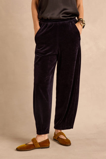 A person stands with hands in pockets, wearing the Night Velvet Jersey Bubble Trousers—a loose black velvet jersey pair with a bubble silhouette—paired with a dark top, yellow-brown velvet loafers, and bracelets, against a beige background.