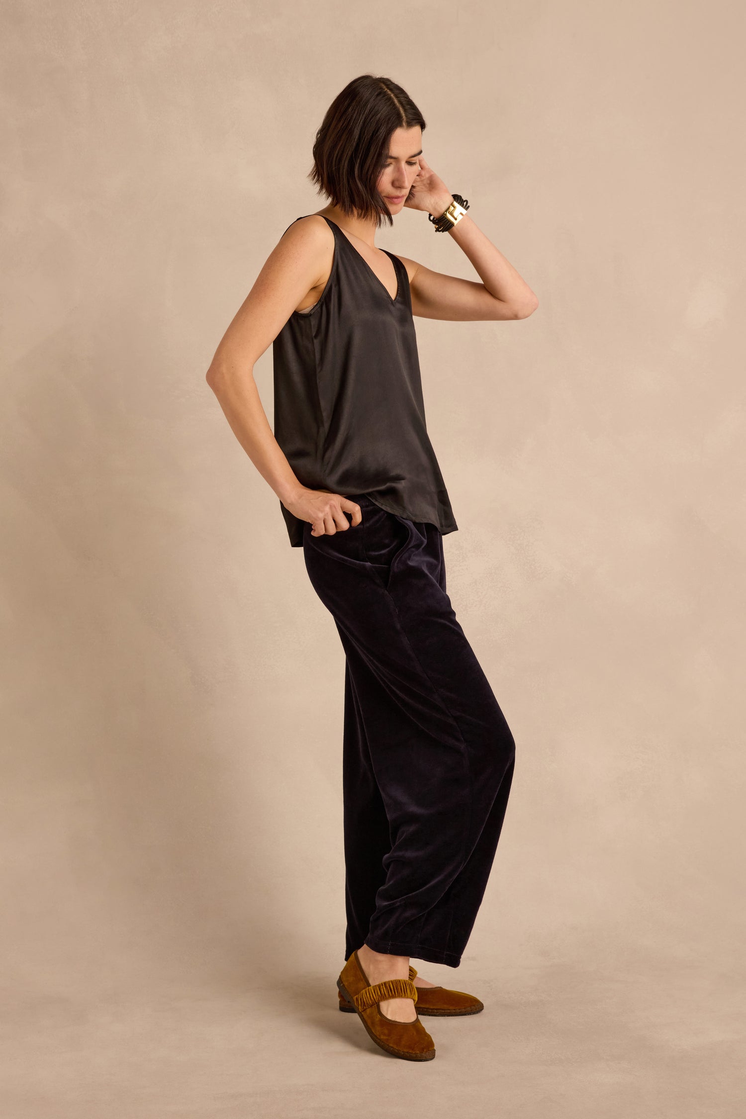 A woman stands sideways against a beige background, wearing a black sleeveless top, the Night Velvet Jersey Bubble Trousers in a loose silhouette, brown moccasin shoes, and a bracelet. All garments are made in England.