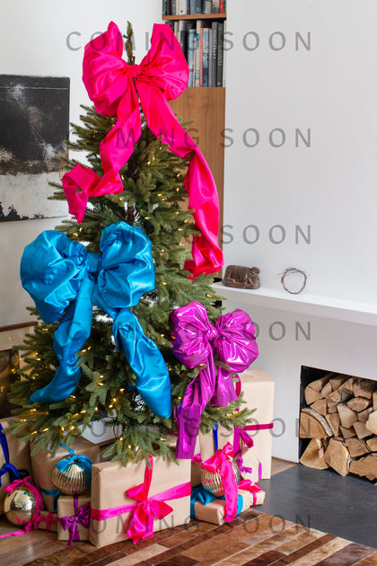 A Christmas tree with bright pink, blue, and purple bows stands among presents in a modern living room, where Velvet Jersey Bubble Trousers are stylishly draped over a chair.