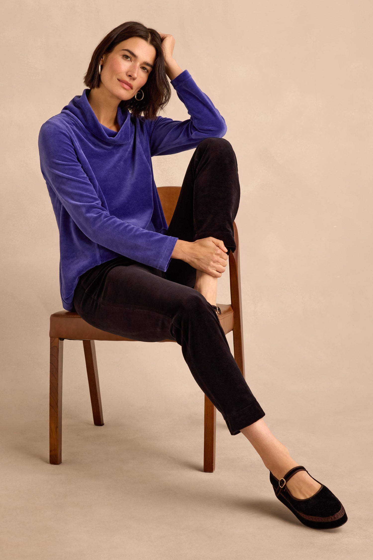 A woman sits on a wooden chair, dressed in a blue long-sleeve top and the Black Velvet Jersey Slim Trousers, which create a streamlined silhouette. She pairs the look with black flat shoes, crossing one leg over the other and resting her hand on her head.