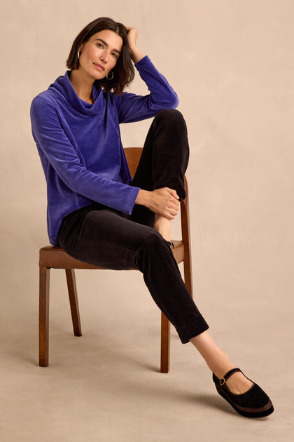 A woman sits on a wooden chair, dressed in a blue long-sleeve top and the Black Velvet Jersey Slim Trousers, which create a streamlined silhouette. She pairs the look with black flat shoes, crossing one leg over the other and resting her hand on her head.