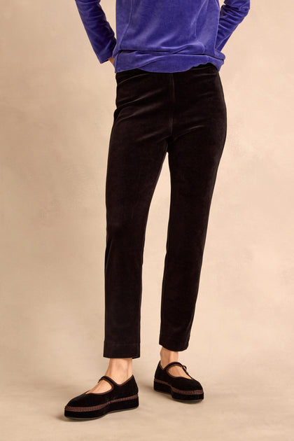 Wearing the Black Velvet Jersey Slim Trousers for a slim silhouette, paired with black Mary Jane shoes and a long-sleeved blue top, this person stands against a neutral background with hands in their pockets.