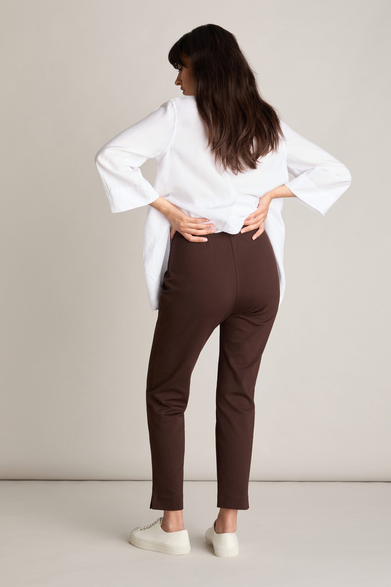 A person with long dark hair faces away, wearing a white top, Ponte Slim Leg Trouser with an elasticated waistband, and white shoes, standing against a light-colored backdrop.