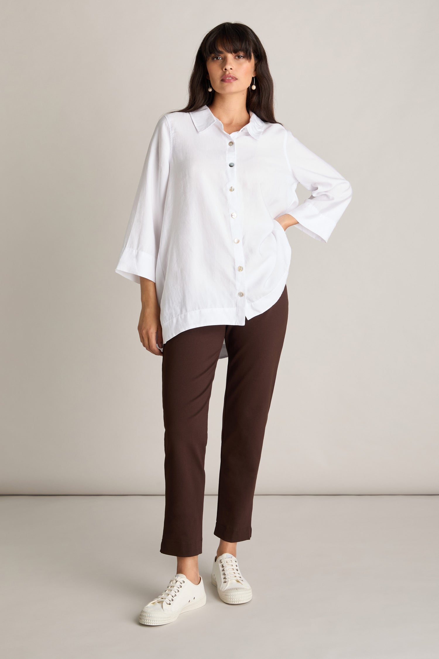 A person stands against a plain backdrop, wearing a long white button-up shirt, Ponte Slim Leg Trousers with an elasticated waistband, and white sneakers.