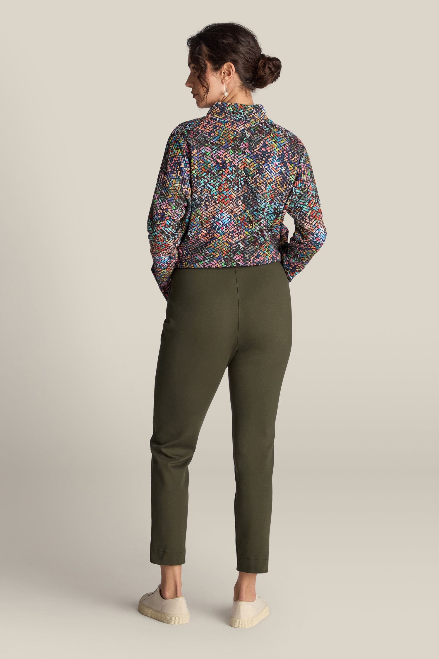 A person stands facing away, showcasing a colorful, patterned top paired with olive green Ponte Slim Leg Trousers that have a tailored touch and an elasticated waistband, completed by light-colored shoes.