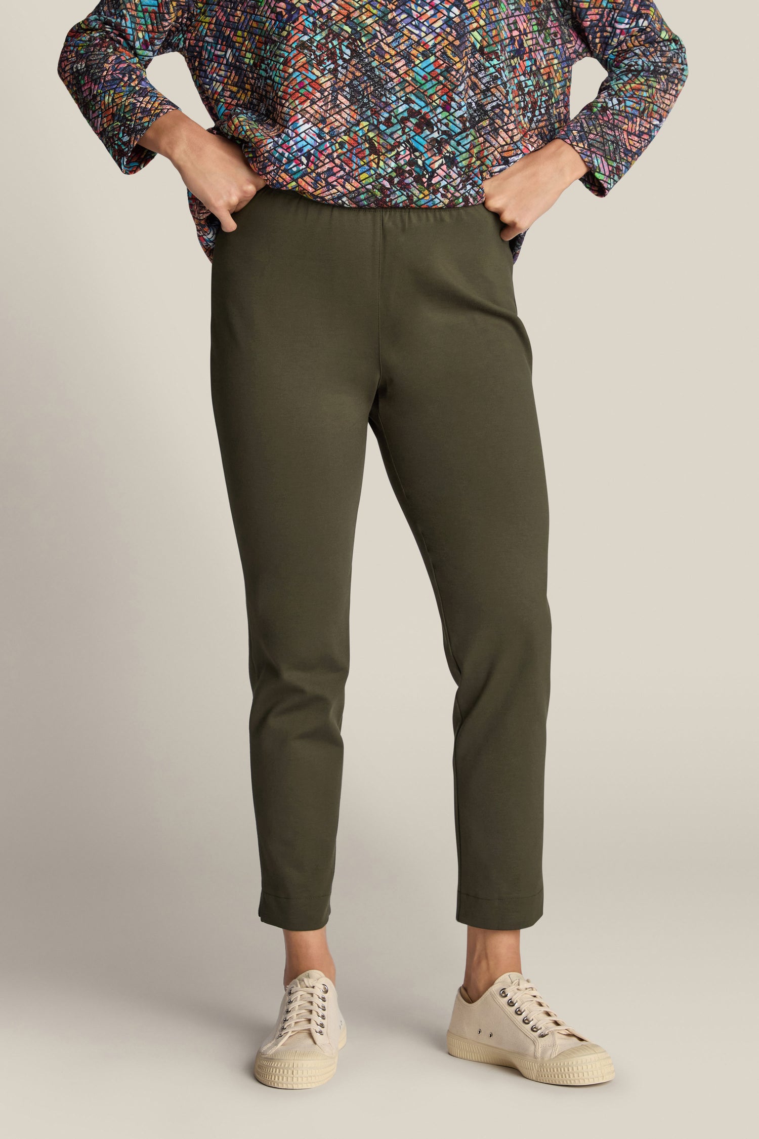 A person stands with hands on hips, wearing olive green Ponte Slim Leg Trousers that add a tailored touch, a colorful long-sleeved top, and beige sneakers. The photo is cropped above the shoulders.