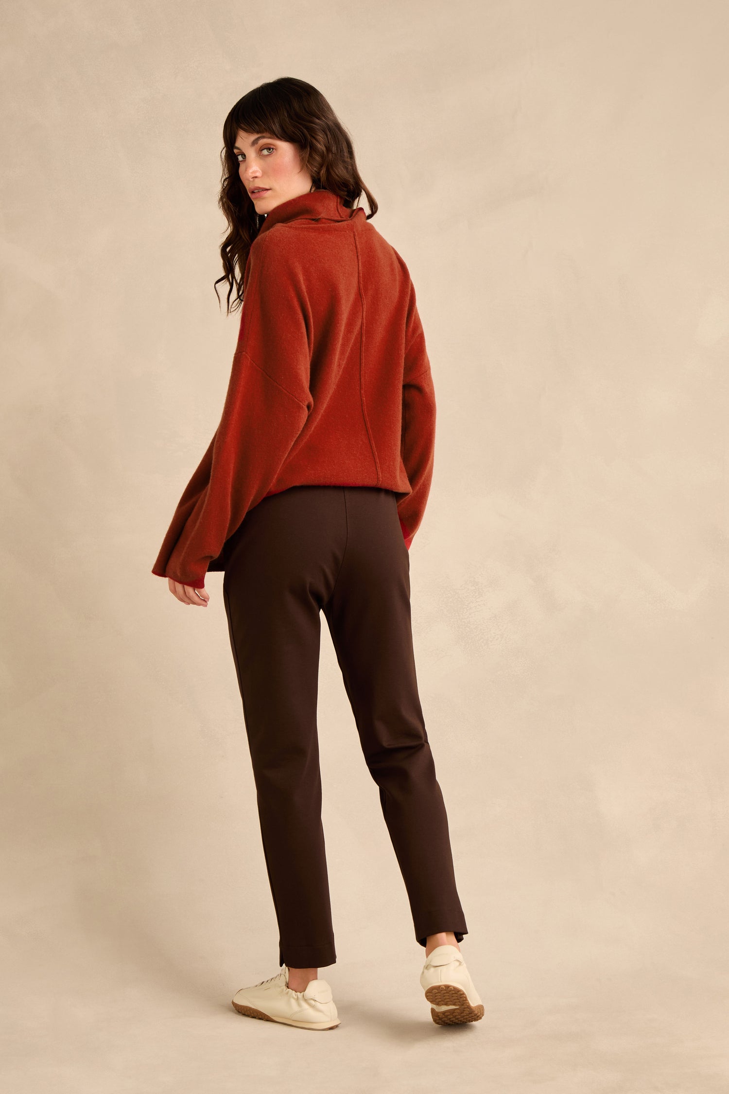 A person stands looking over their shoulder, dressed in a rust-colored sweater and Chocolate Ponte Slim Leg Trousers with a pull-on elasticated waist, paired with white shoes against a plain beige background.