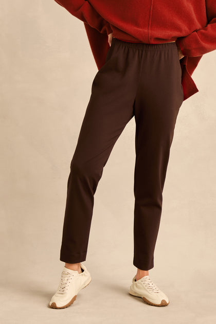 A person wearing a red top, the Chocolate Ponte Slim Leg Trousers with a pull-on elasticated waist, and white sneakers stands with hands in pockets against a plain beige background.