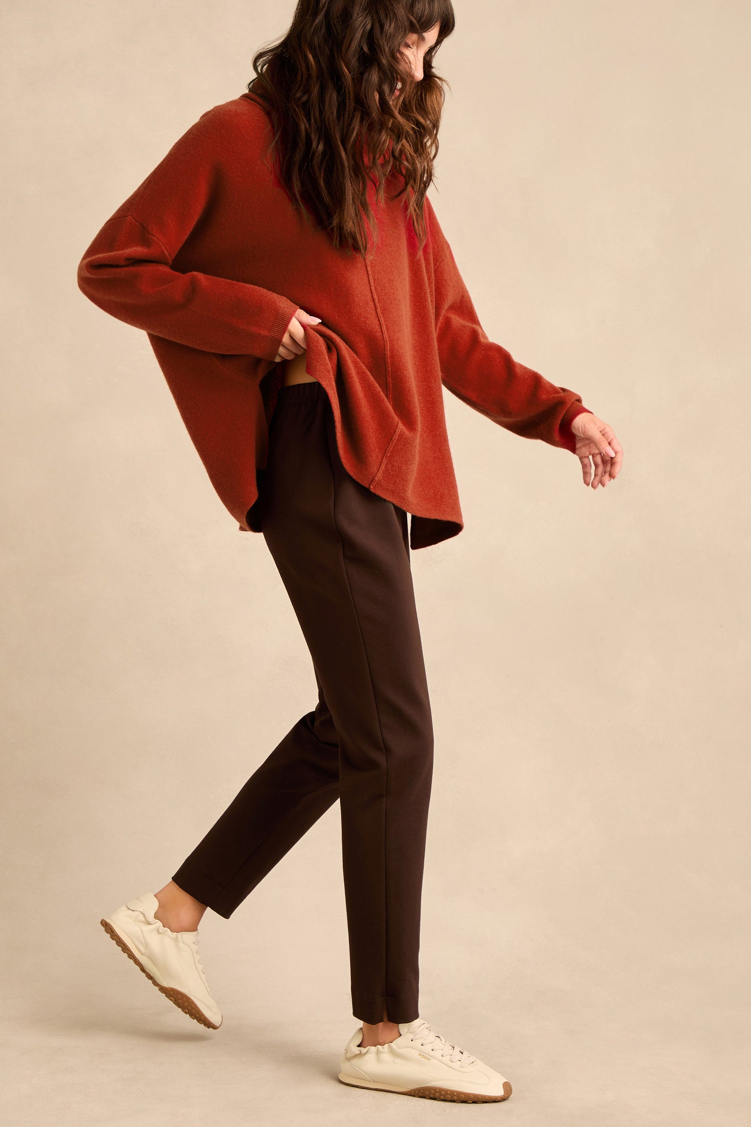 Woman standing sideways, wearing a loose rust-colored sweater, Chocolate Ponte Slim Leg Trousers with a pull-on elasticated waist, and white sneakers against a neutral background. She has long, wavy brown hair and is mid-step.