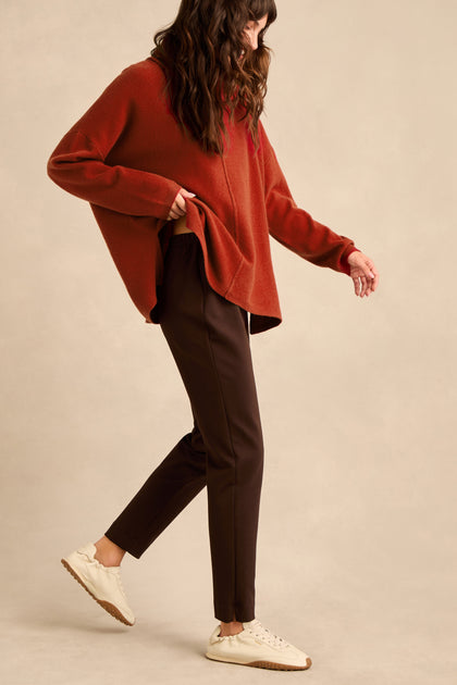 Woman standing sideways, wearing a loose rust-colored sweater, Chocolate Ponte Slim Leg Trousers with a pull-on elasticated waist, and white sneakers against a neutral background. She has long, wavy brown hair and is mid-step.