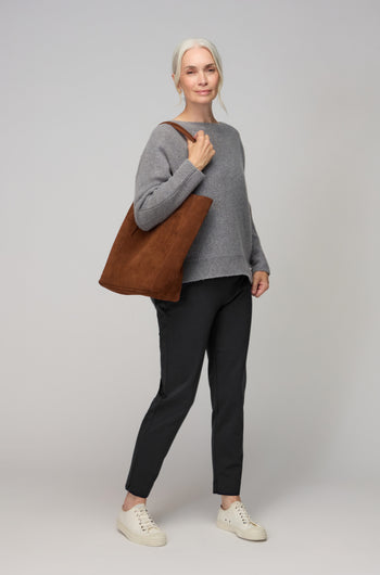 An older woman with gray hair in a ponytail, wearing a gray sweater, Ponte Slim Leg Trousers, white sneakers, and carrying a brown tote bag stands against a plain light background.