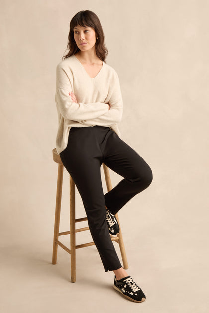 A person sits on a wooden stool against a plain beige background, wearing a cream-colored sweater, black sneakers, and the Black Ponte Slim Leg Trouser.