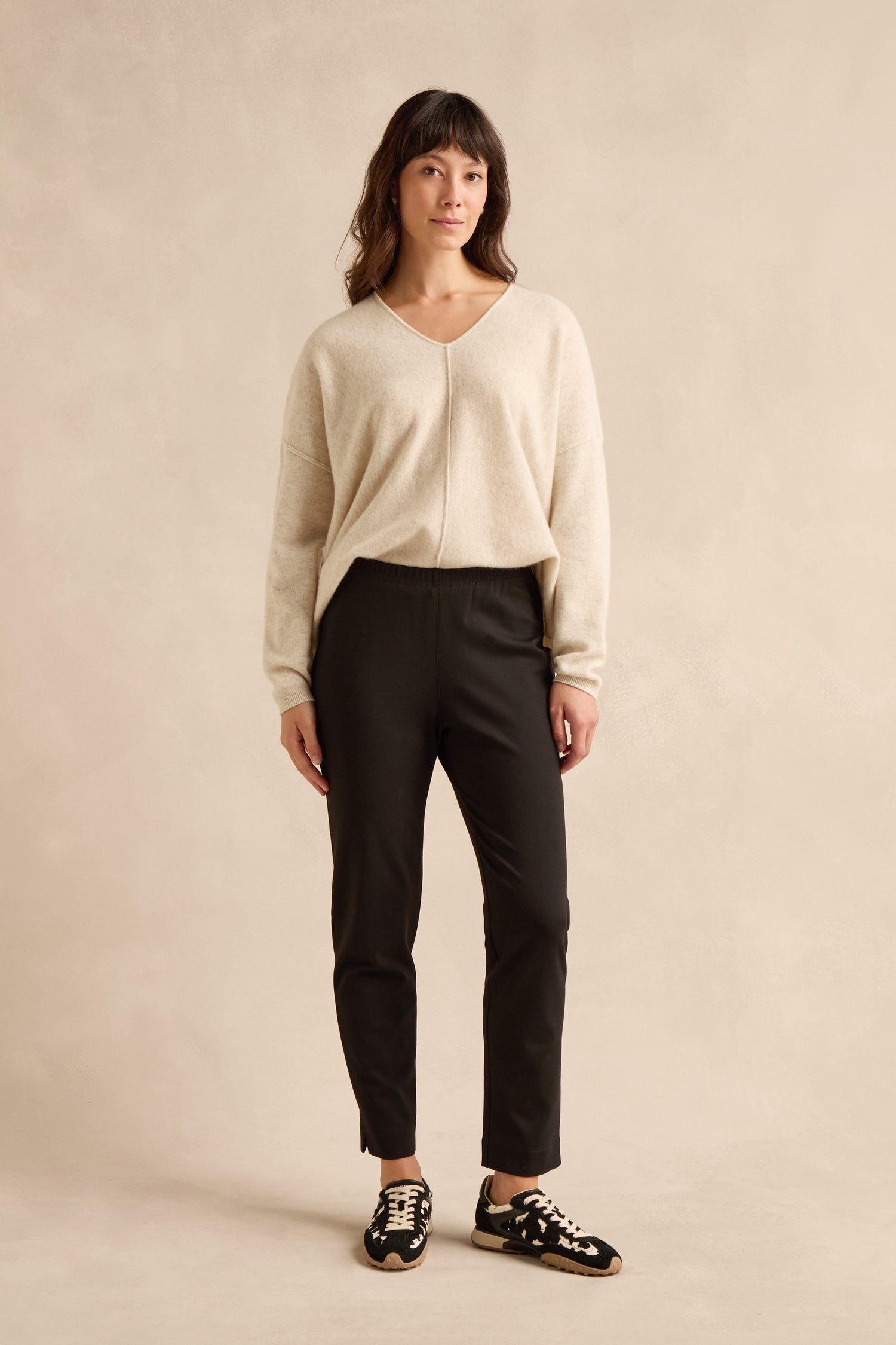 A woman stands facing forward, wearing the Black Ponte Slim Leg Trouser along with a beige long-sleeve sweater and black sneakers with white laces against a neutral background.