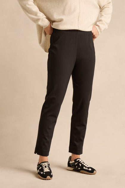 A person wearing Black Ponte Slim Leg Trousers, a cream sweater, and black sneakers with white laces stands with hands in their pockets against a neutral background.