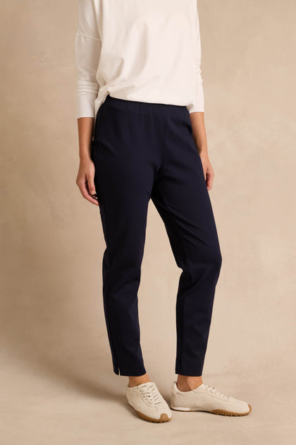 A person wearing a white long-sleeve top, the Midnight Ponte Slim Leg Trouser in navy with an elasticated waist, and beige sneakers stands against a plain beige background.