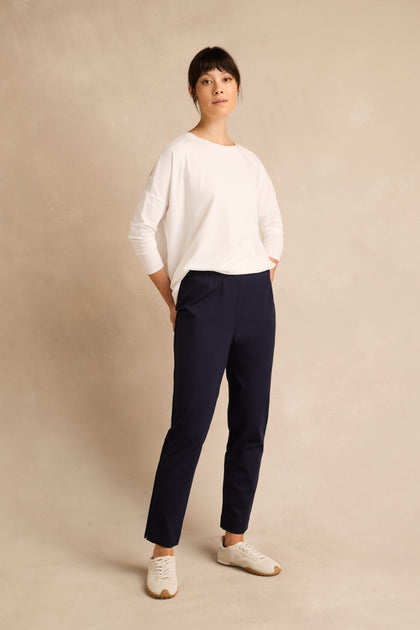 Person stands against a light beige background, wearing a white long-sleeve shirt, the Midnight Ponte Slim Leg Trouser in navy, and white sneakers, with hands in pockets and a neutral expression.