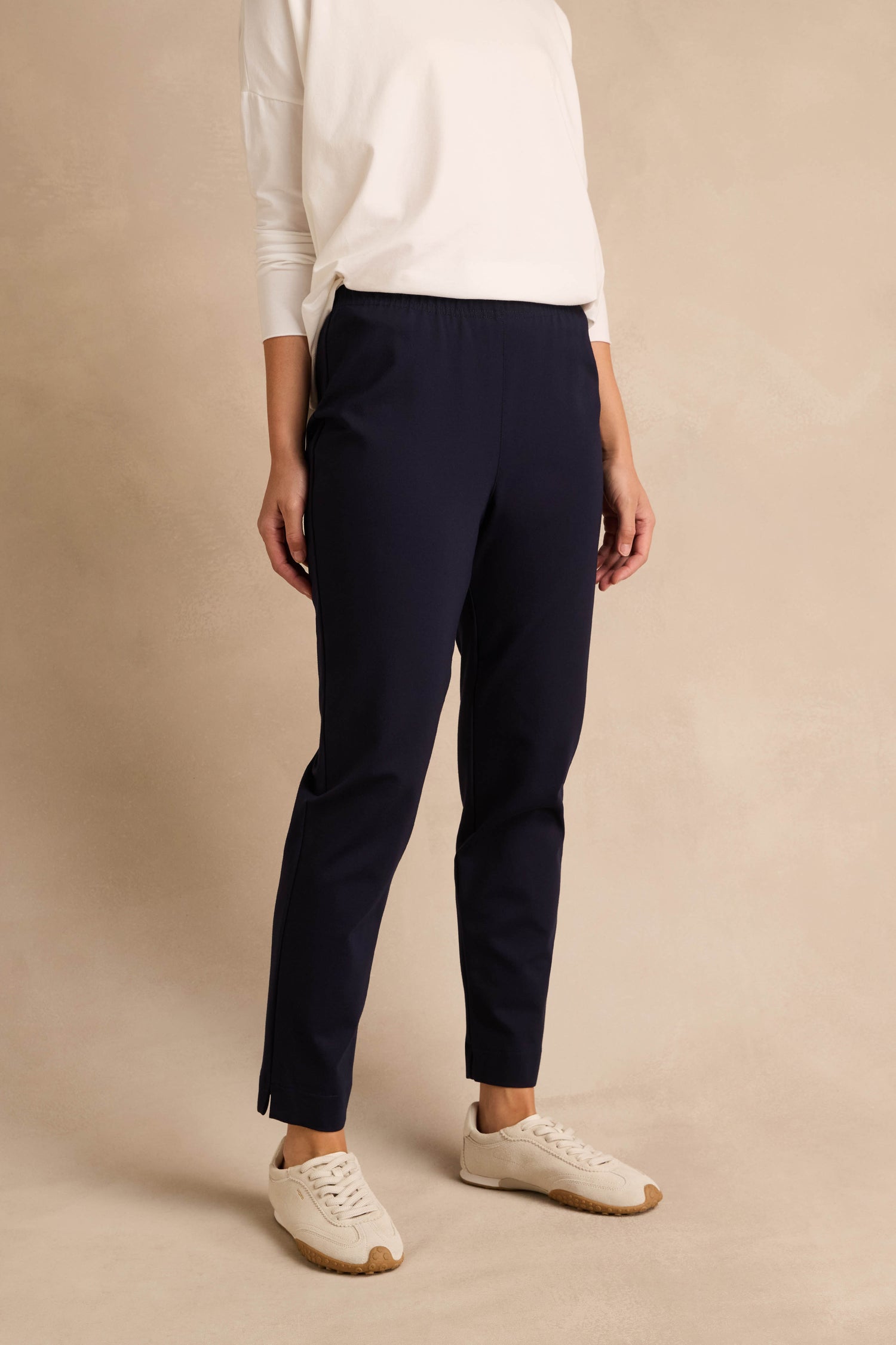 Person wearing a white long-sleeve top, Midnight Ponte Slim Leg Trouser with an elasticated waist, and white sneakers, standing against a beige background.