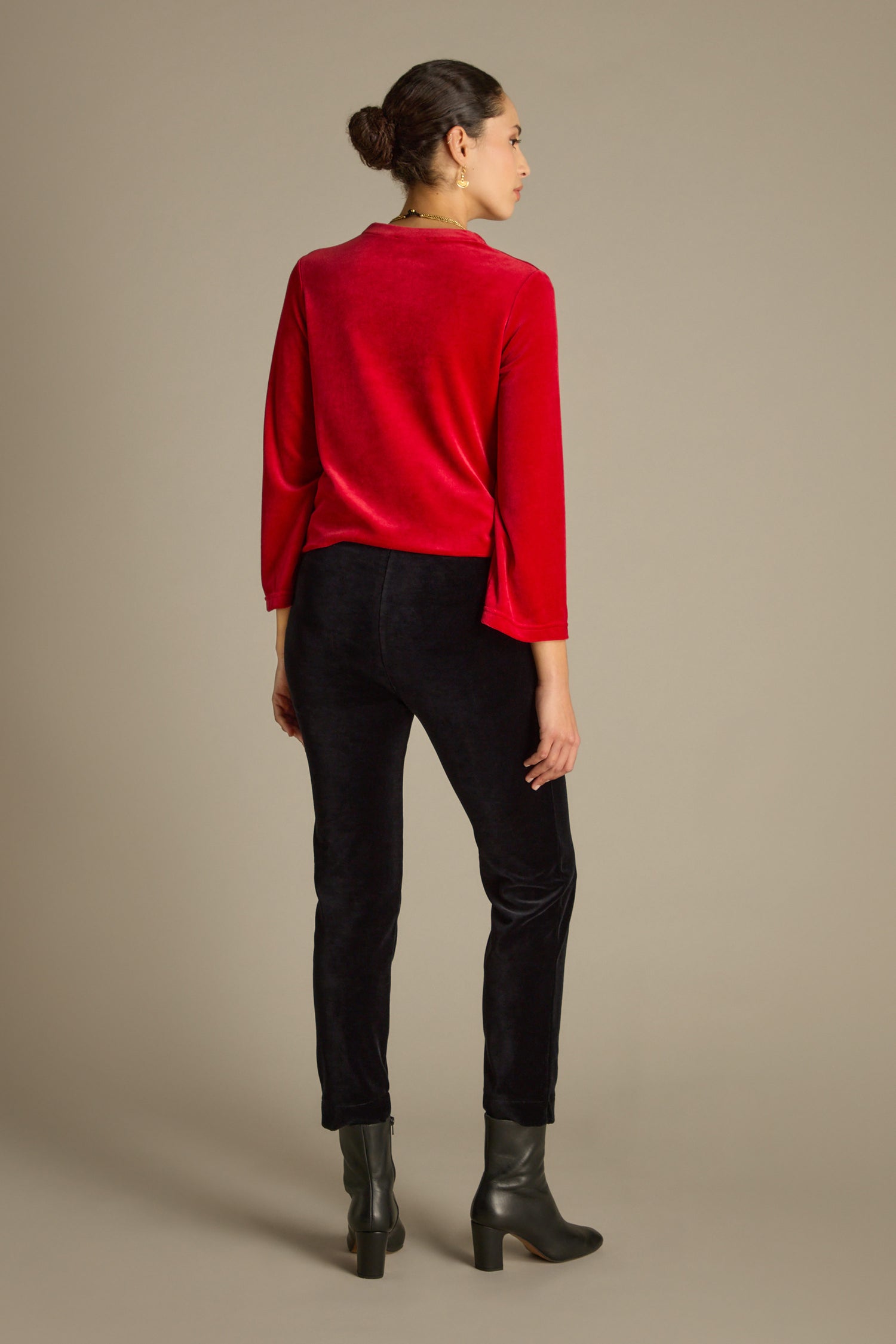 A person stands facing away, wearing a red top, sleek black Velvet Jersey Slim Leg Trousers with an elasticated waist, and black boots against a neutral background.