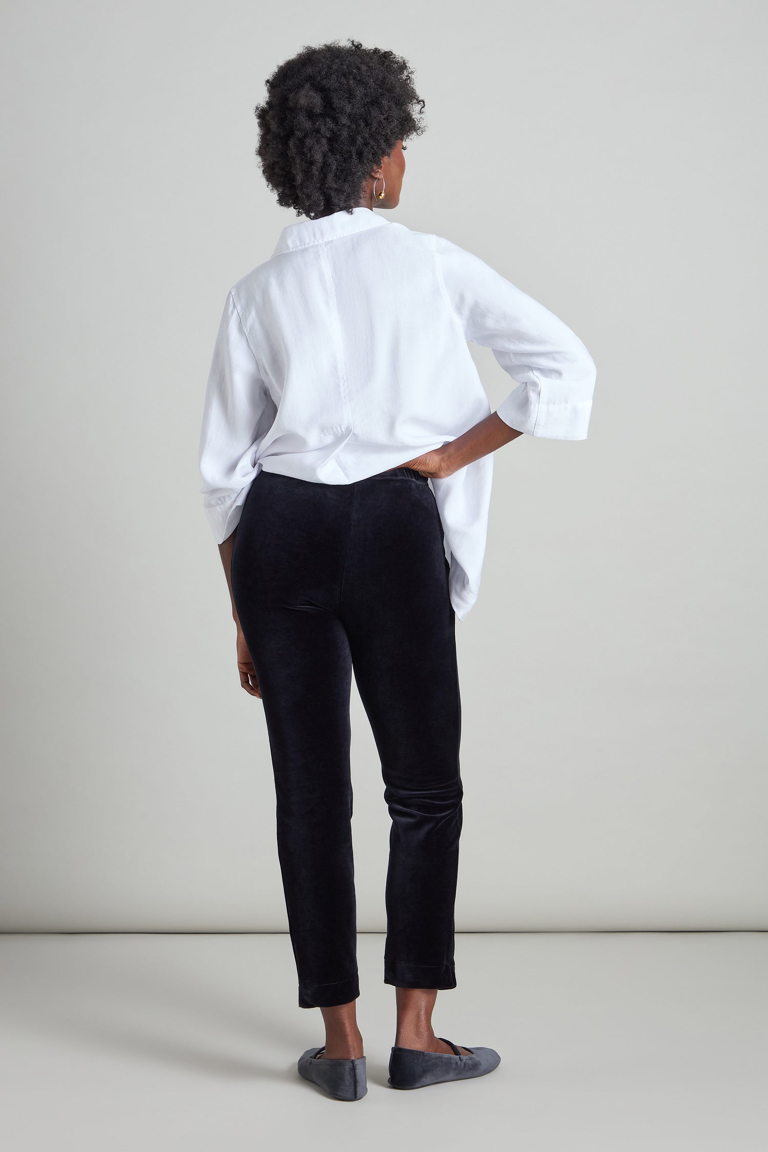 A person stands facing away, wearing a white shirt, Velvet Jersey Slim Leg Trouser, and gray shoes against a plain background.