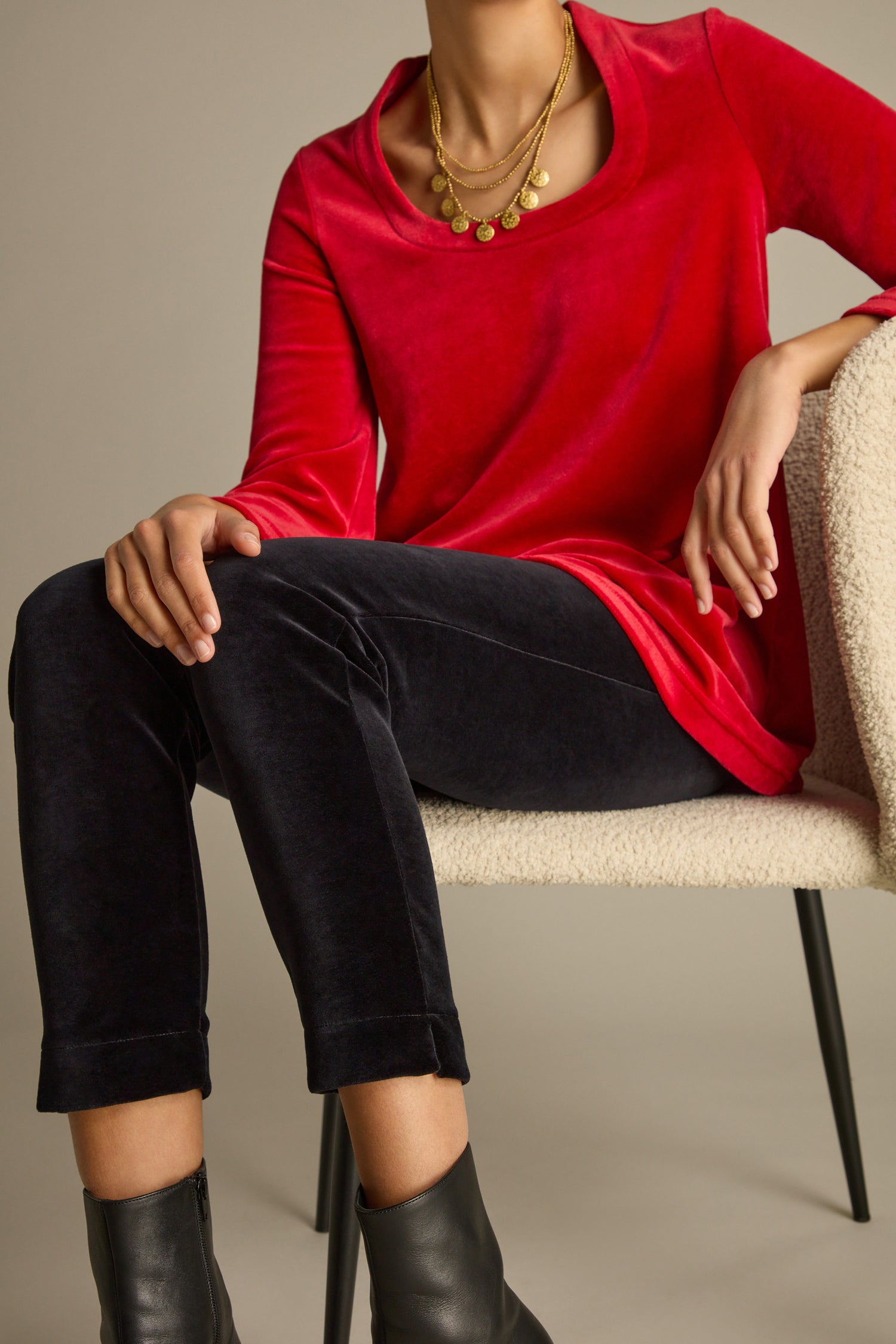 A person sits in a chair, wearing a red long-sleeve top, Velvet Jersey Slim Leg Trousers in black, and black ankle boots, complemented by a layered gold necklace.