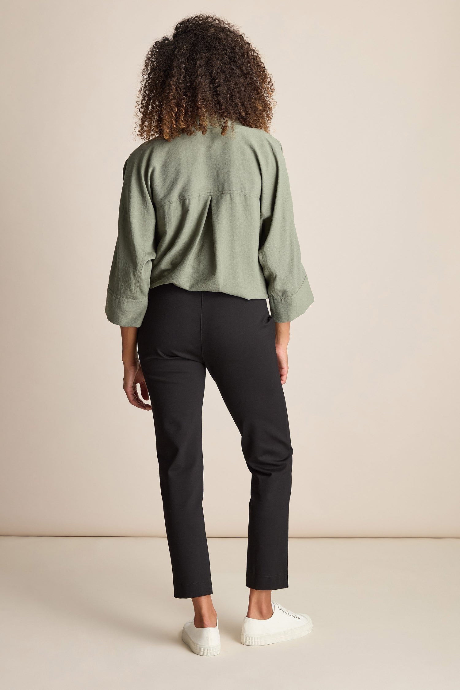 A person with curly hair is standing with their back to the camera, wearing a green long-sleeve blouse, Ponte Slim Leg Trousers, and white sneakers.