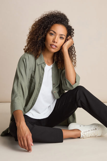 A person with curly hair is sitting on the floor, wearing a green shirt over a white t-shirt, Ponte Slim Leg Trouser in black, and white shoes, with one hand resting on their face.