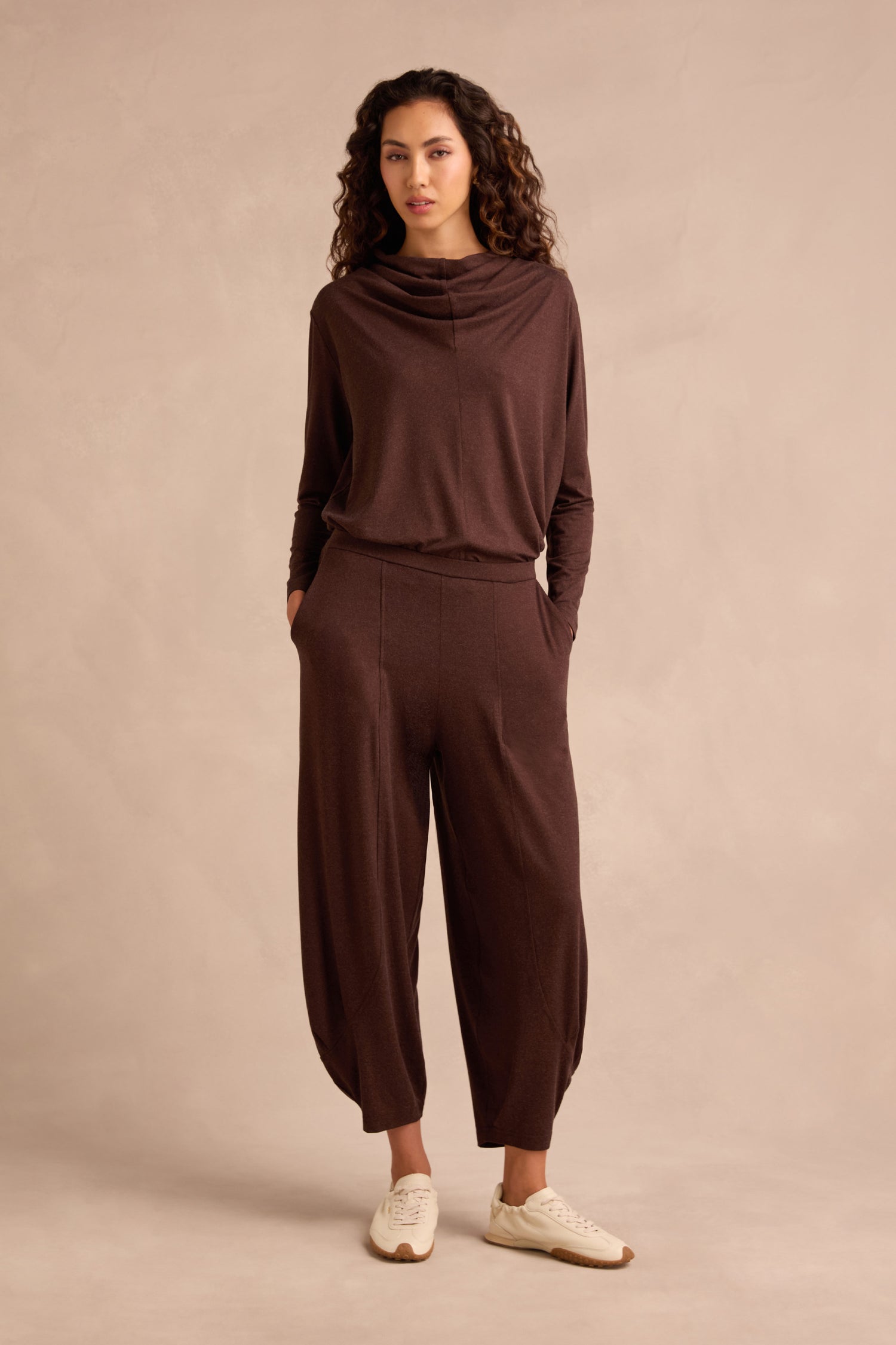 A person stands facing forward, wearing a brown long-sleeve top with a draped collar, Soft Viscose Jersey Bubble Trousers, and white sneakers, against a beige background.