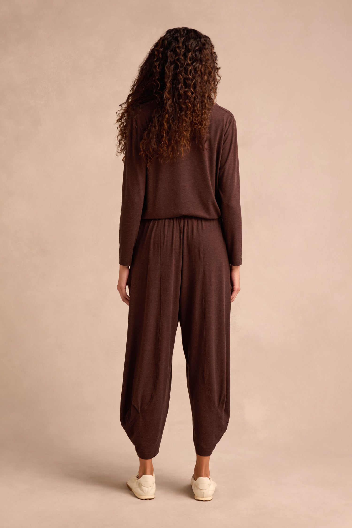 A person with curly hair stands facing away, wearing loose brown long-sleeve top and matching Soft Viscose Jersey Bubble Trouser, paired with white shoes, against a plain beige background.