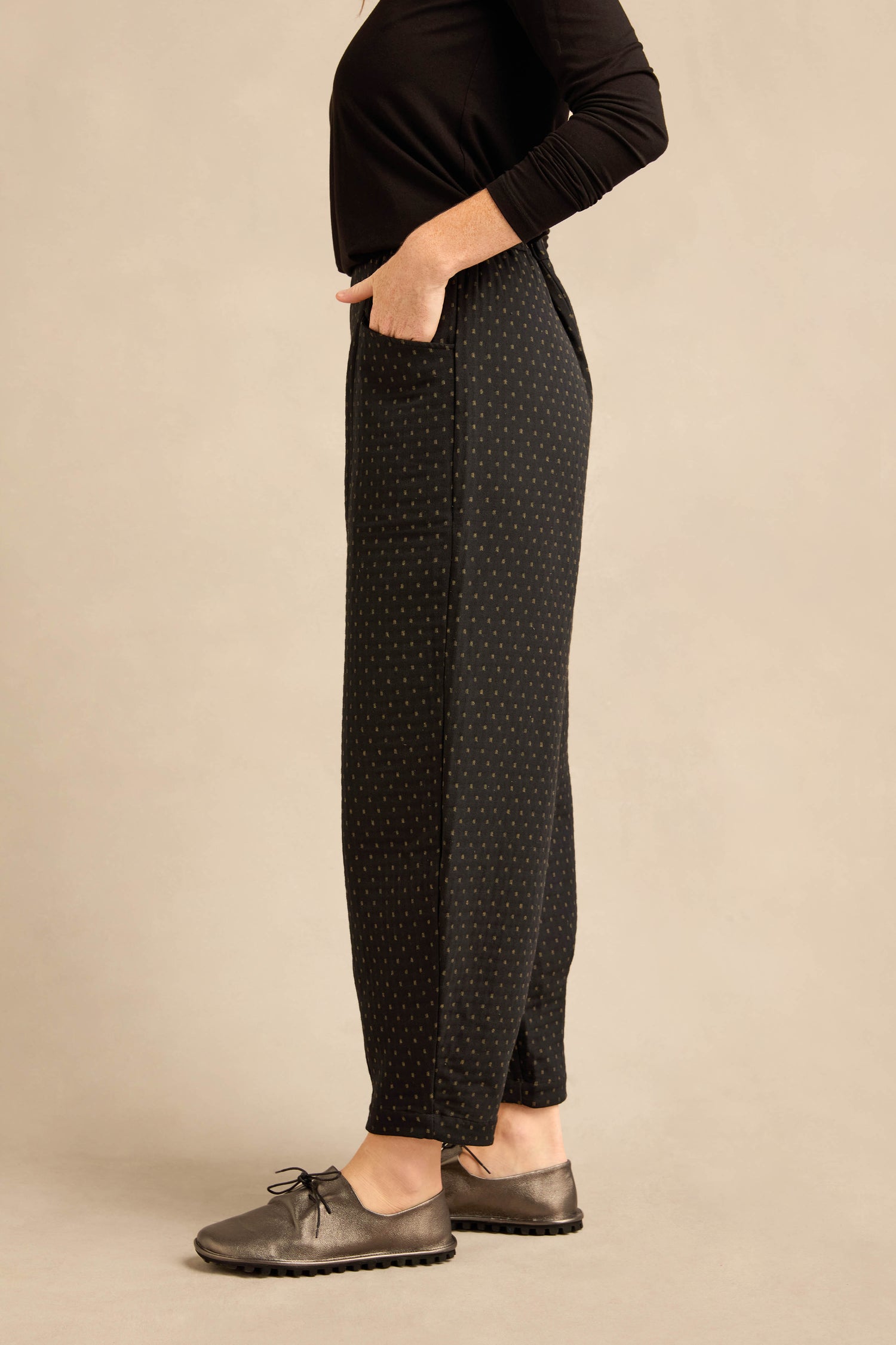 A person stands sideways against a plain beige background, wearing a black long-sleeve top, gray lace-up shoes, and Square Jacquard Bubble Trousers crafted from a cotton-rich blend with pockets.