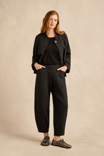 A woman stands with her hands in her pockets, wearing Square Jacquard Bubble Trousers featuring a cotton-rich blend and subtle dot pattern, paired with a black jacket, black t-shirt, and black shoes against a neutral background.