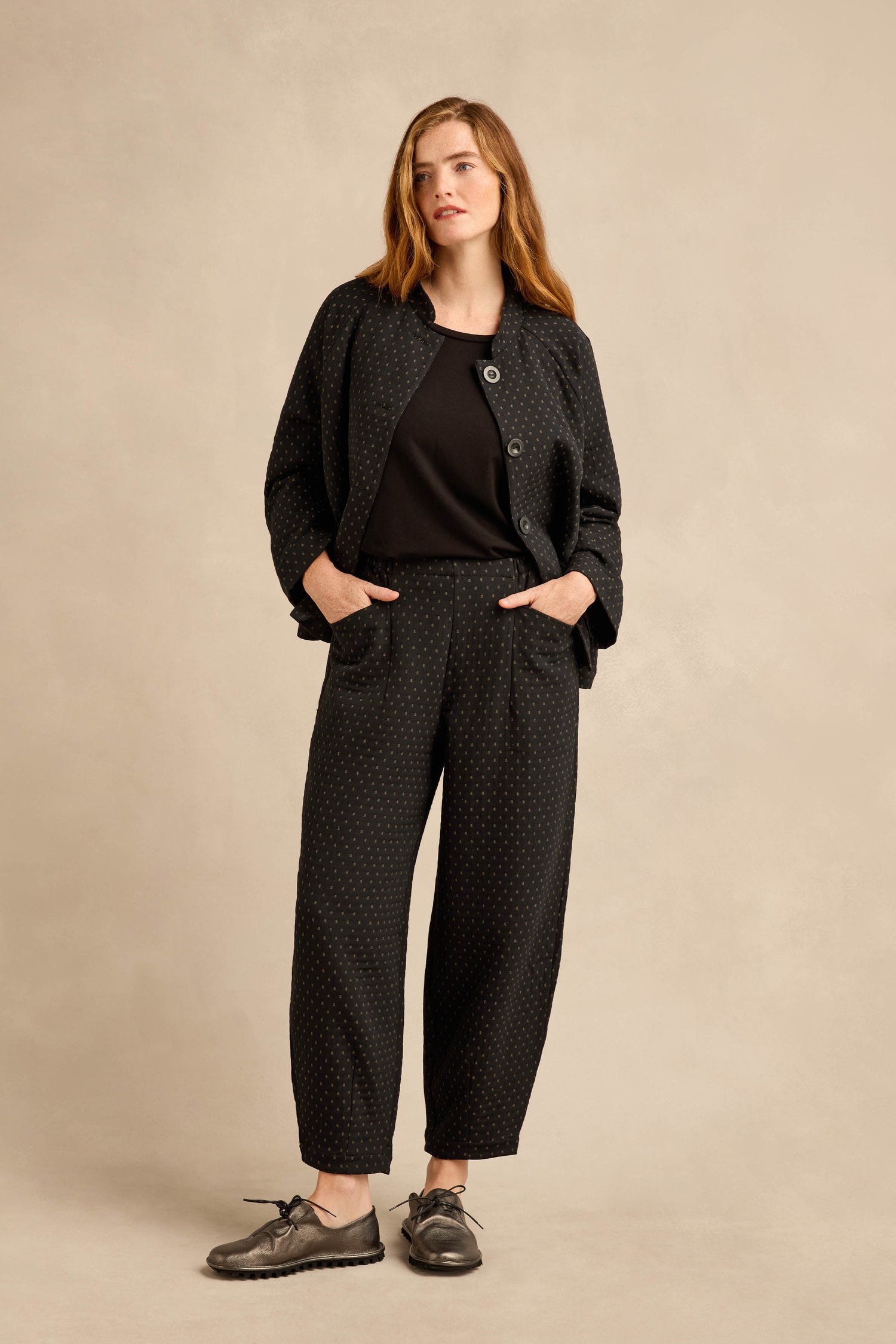 A woman stands facing forward against a plain beige background, wearing Square Jacquard Bubble Trousers with a matching black patterned jacket, a black shirt, and gray lace-up shoes. Her hands are in her pockets.
