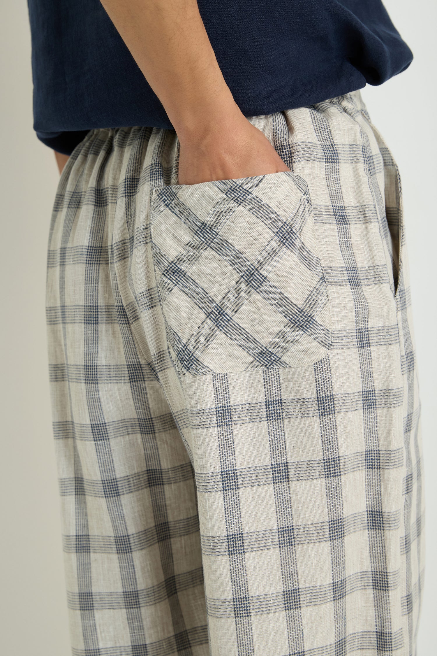 A hand rests in the pocket of Grid Check Linen Bubble Trouser, featuring a beige and blue check pattern with a signature bubble shape; only the lower torso and upper legs are visible.