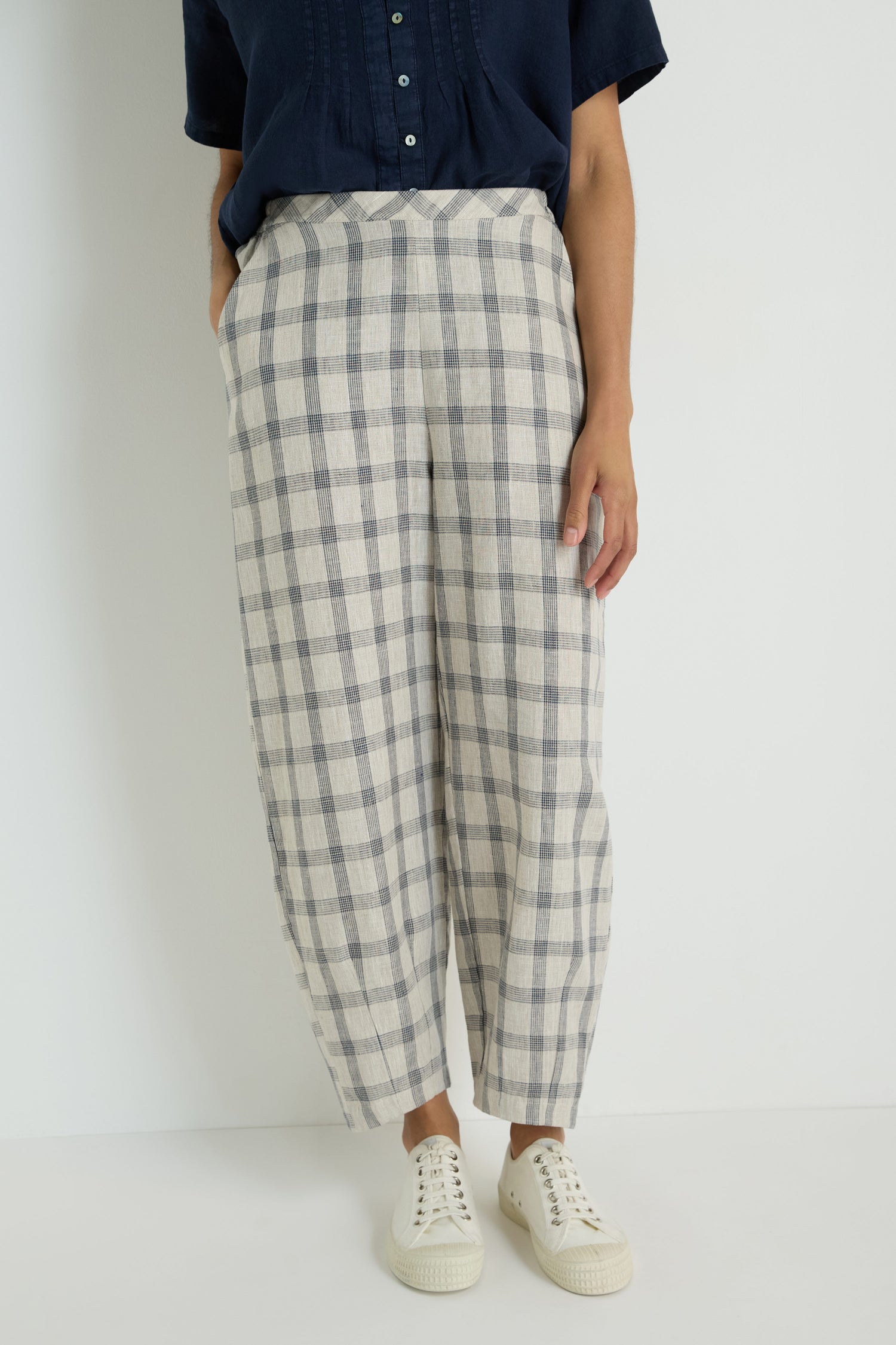 A woman faces away against a white background, wearing a short-sleeve beige top, Grid Check Linen Bubble Trouser with its signature bubble shape, and brown slip-on shoes.