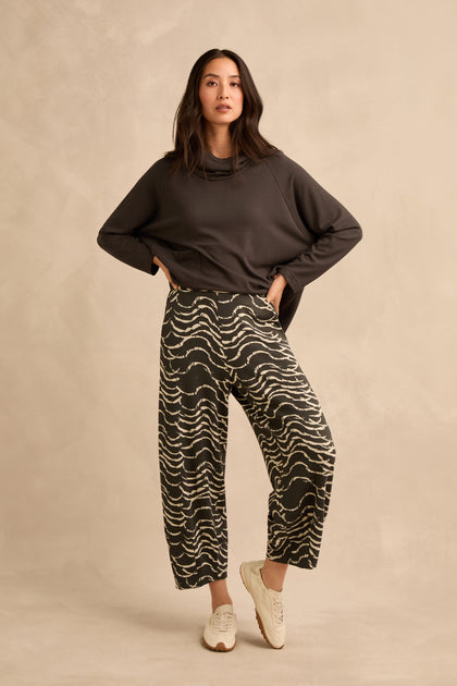 A woman stands against a beige background wearing a dark long-sleeve top, white sneakers, and the Dancing Waves Jersey Bubble Trousers, featuring a stretch jacquard wide-leg print. She looks directly at the camera with one hand on her hip.
