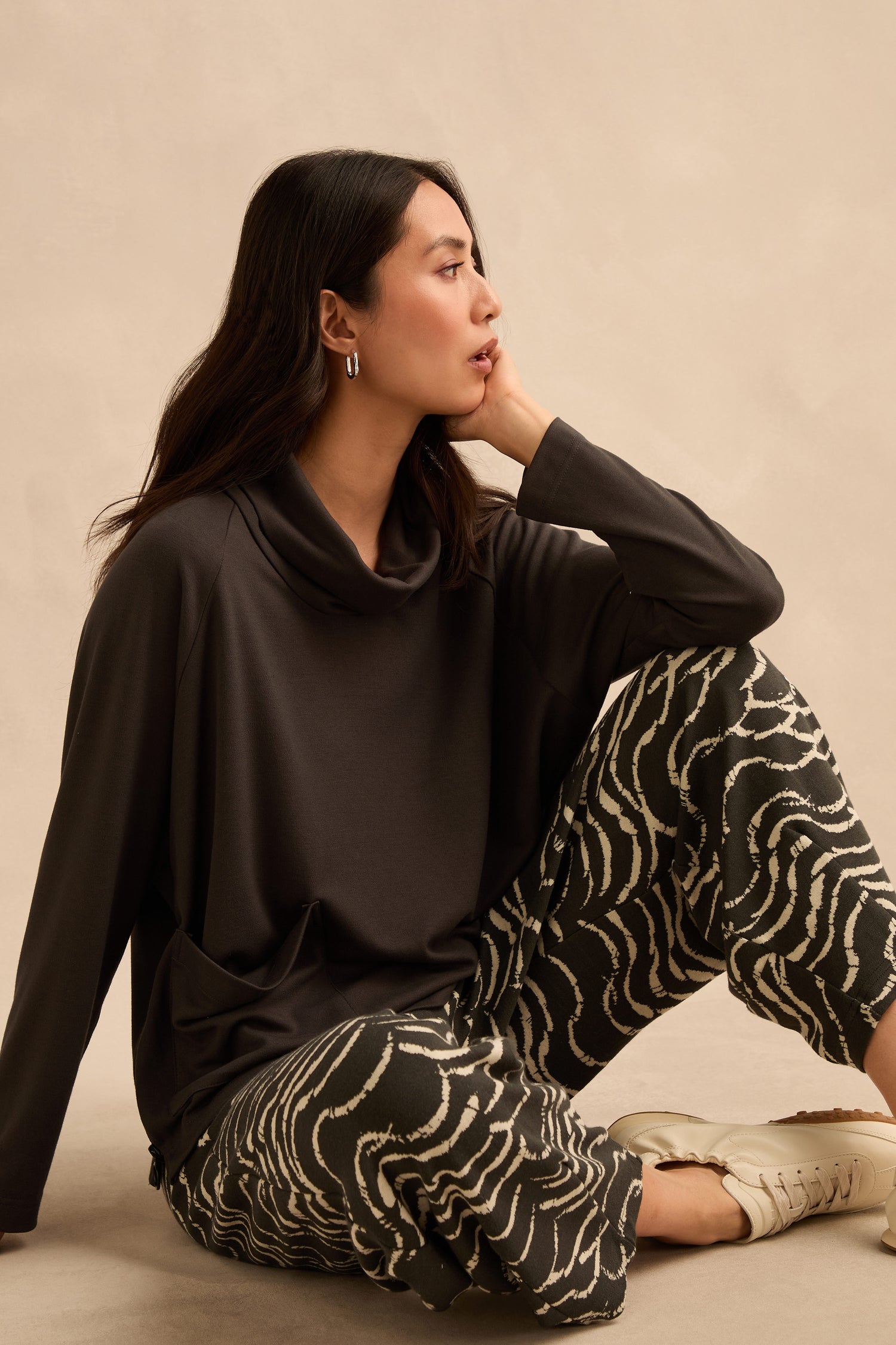 A woman sits on the floor against a neutral background, looking to the left. She wears a dark cowl-neck top with Dancing Waves Jersey Bubble Trousers and crisp white sneakers.