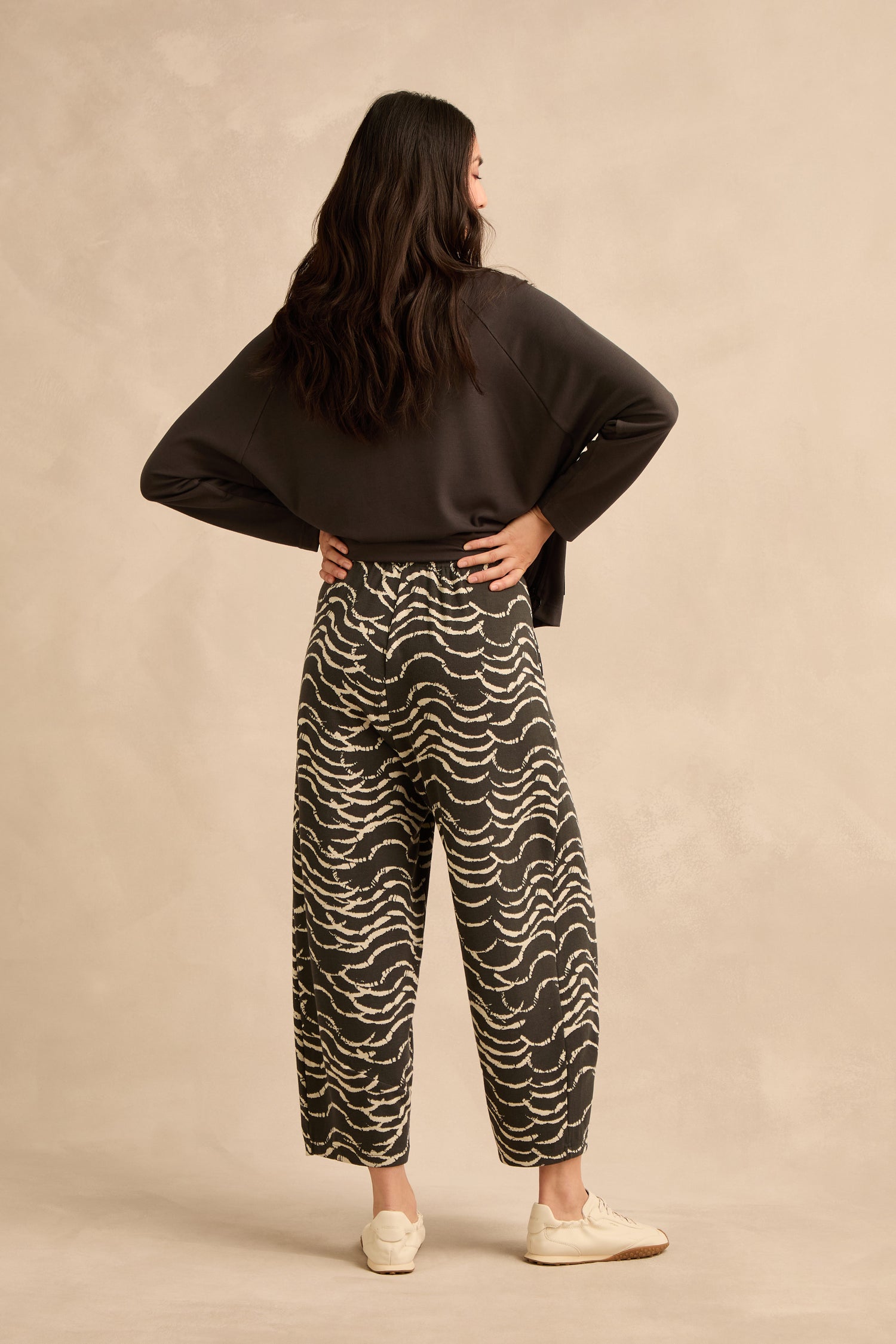 A woman stands with her back to the camera, dressed in a dark sweatshirt, Dancing Waves Jersey Bubble Trousers, and white sneakers, set against a neutral, textured background.