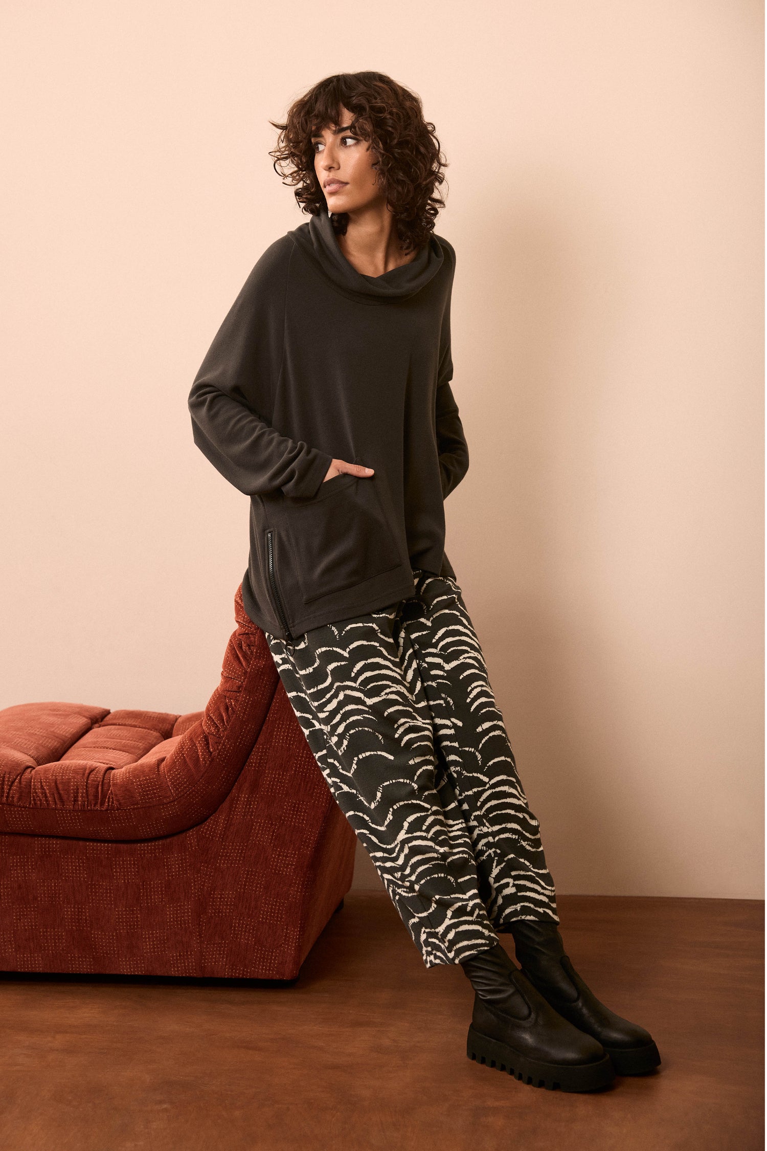 Wearing Etched Wave Jersey Bubble Trousers with a dark cowl-neck sweater and black boots, a person leans against a rust-colored chaise lounge on a brown floor with a beige wall.